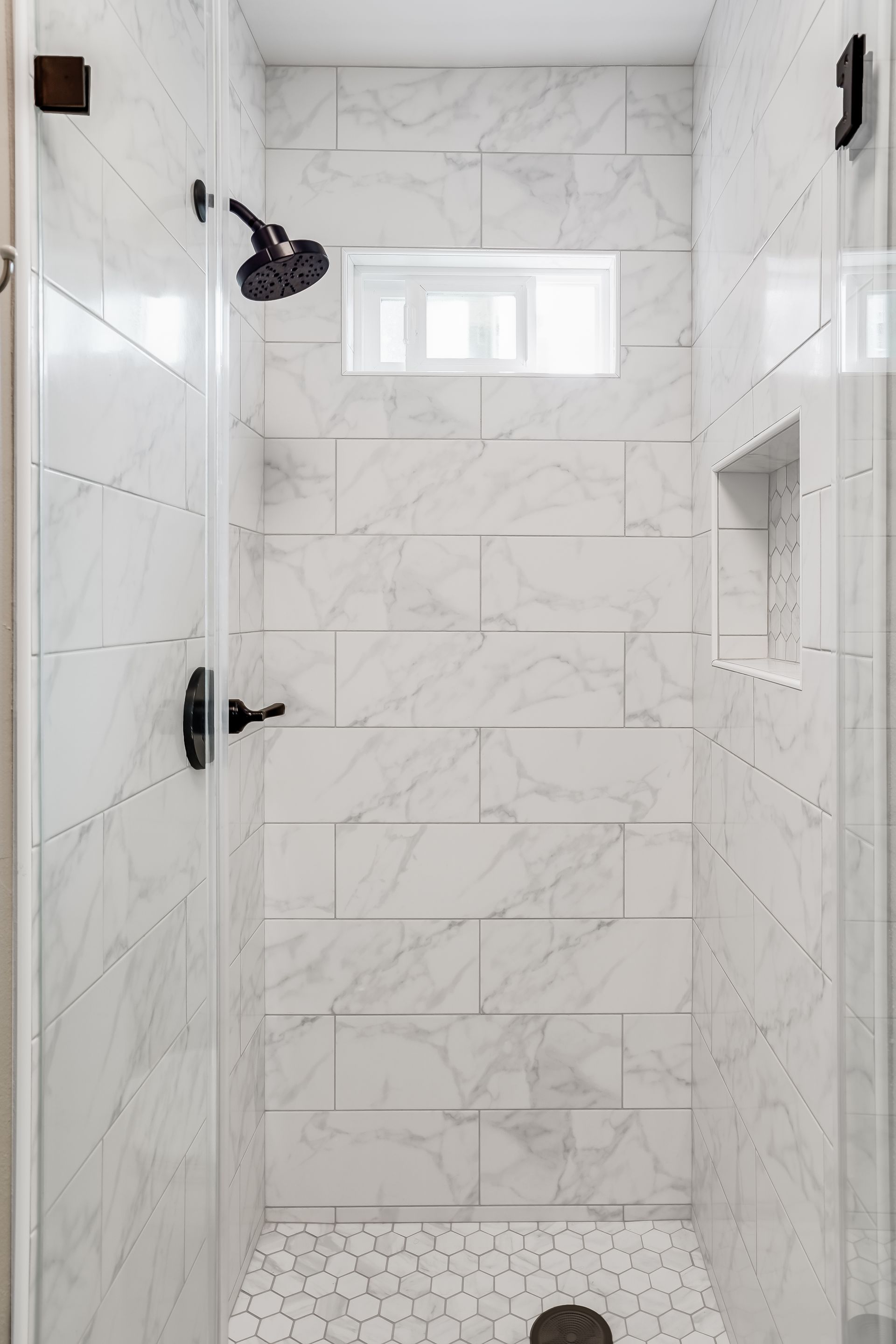 White tiled shower with bronze fixtures and a small window.