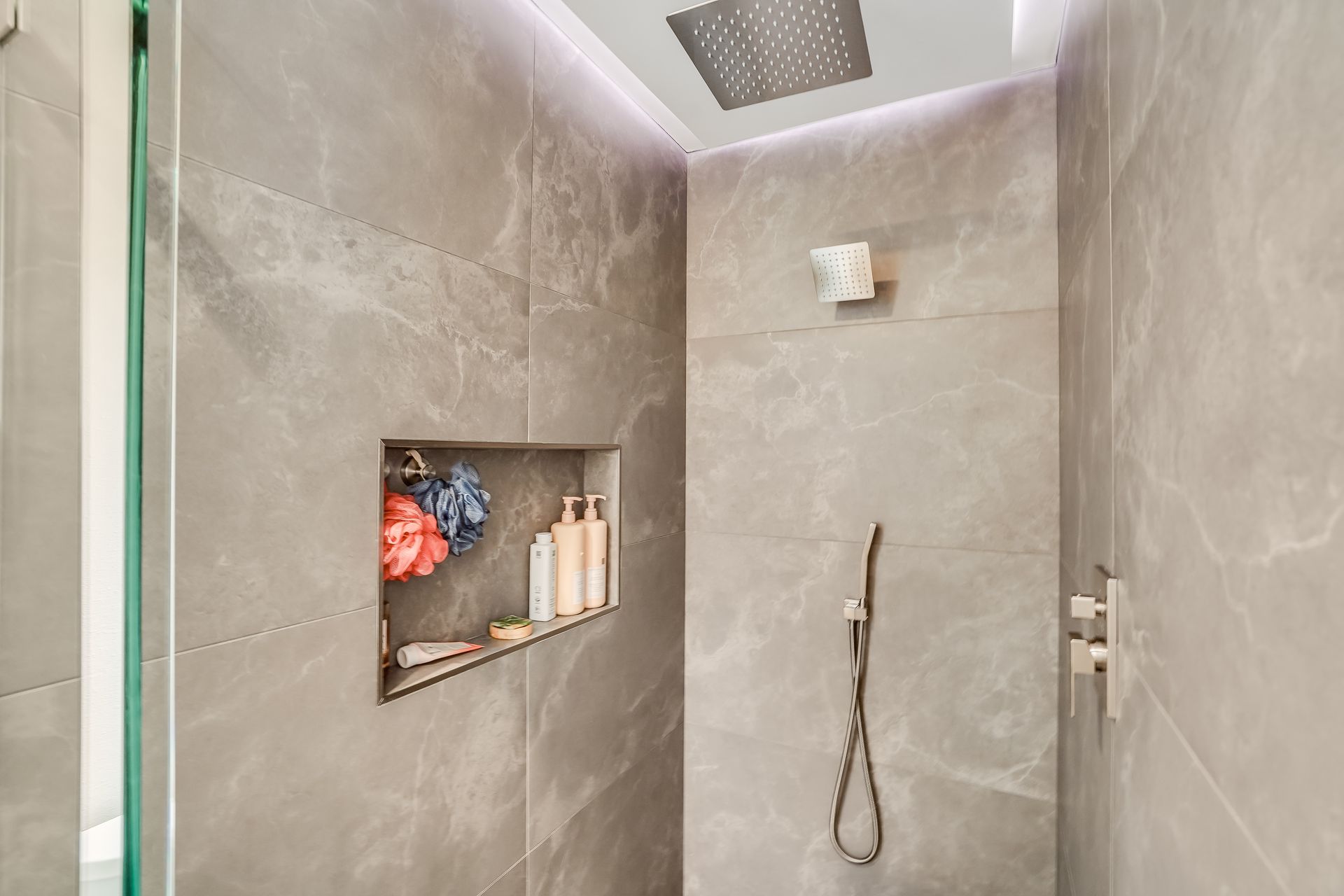 Gray tiled shower with a built-in shelf containing toiletries and a rain shower head.