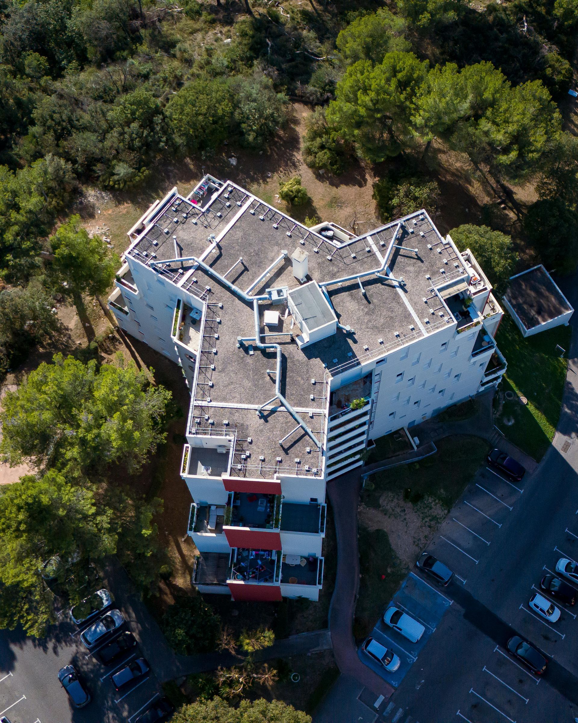 Aerial view of a white multi-story building with a red and gray roof, surrounded by trees and a parking lot.