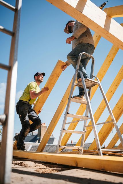 Two construction workers installing wooden beams on a roof, sunny day, one on a ladder.