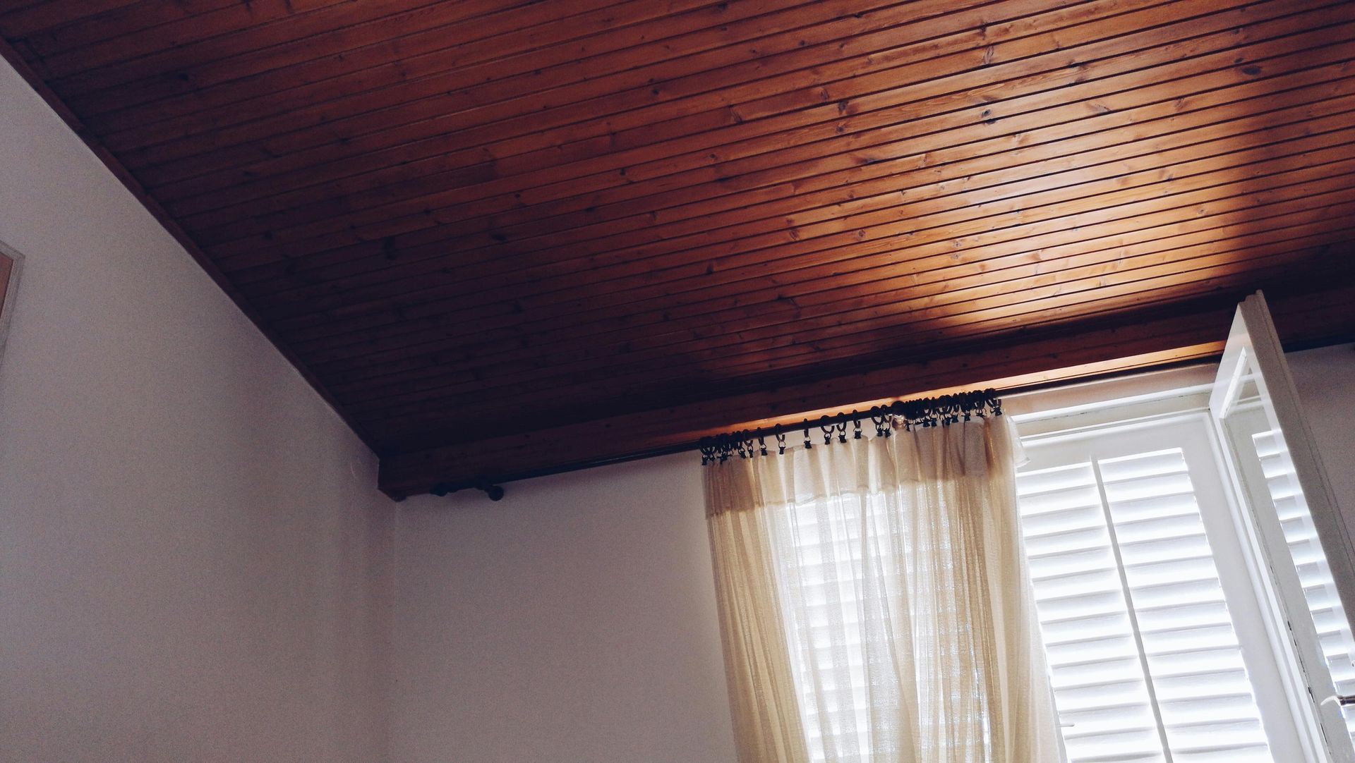 Wooden ceiling with sunlight, window with blinds and beige curtain, white wall.