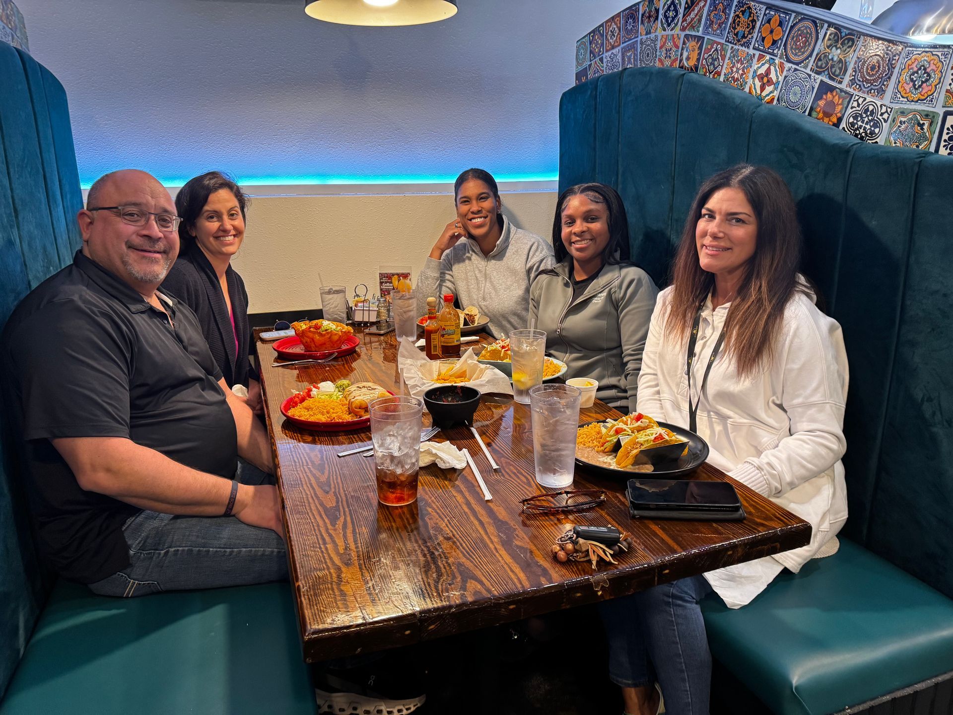 Five people seated around a restaurant booth table with drinks and dishes, smiling during a meal