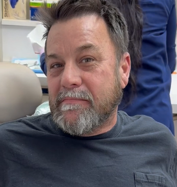 Smiling bearded man seated indoors, wearing a dark shirt with a blue-clad person behind him