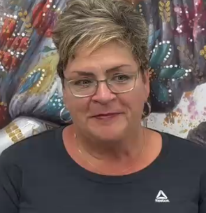 Woman with short blond hair and glasses, wearing a dark shirt, seated before a colorful patterned backdrop.