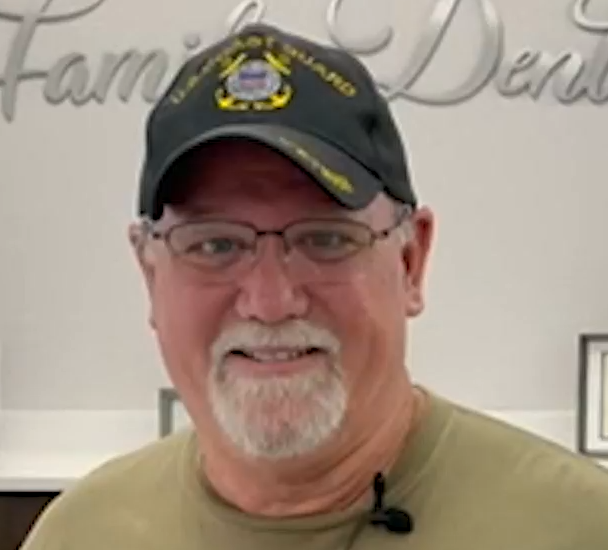 Smiling man wearing glasses and a black cap, standing indoors against a light wall