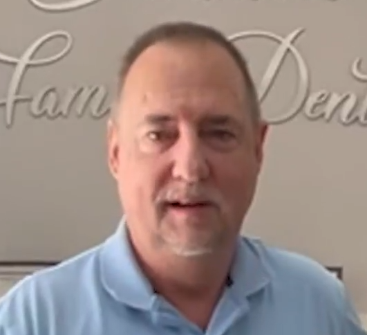 Man in a light blue polo shirt speaking indoors, standing before a beige wall with cursive lettering.
