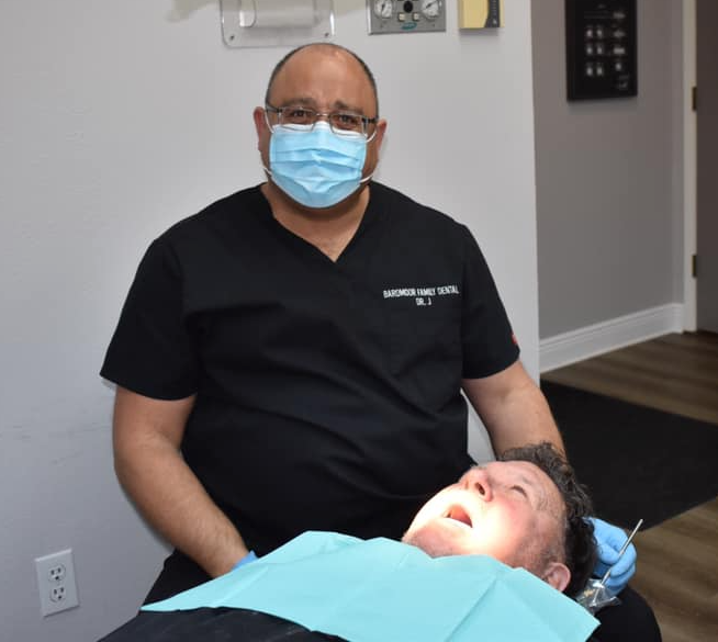 Dentist in black scrubs and mask sits beside patient in dental chair during exam