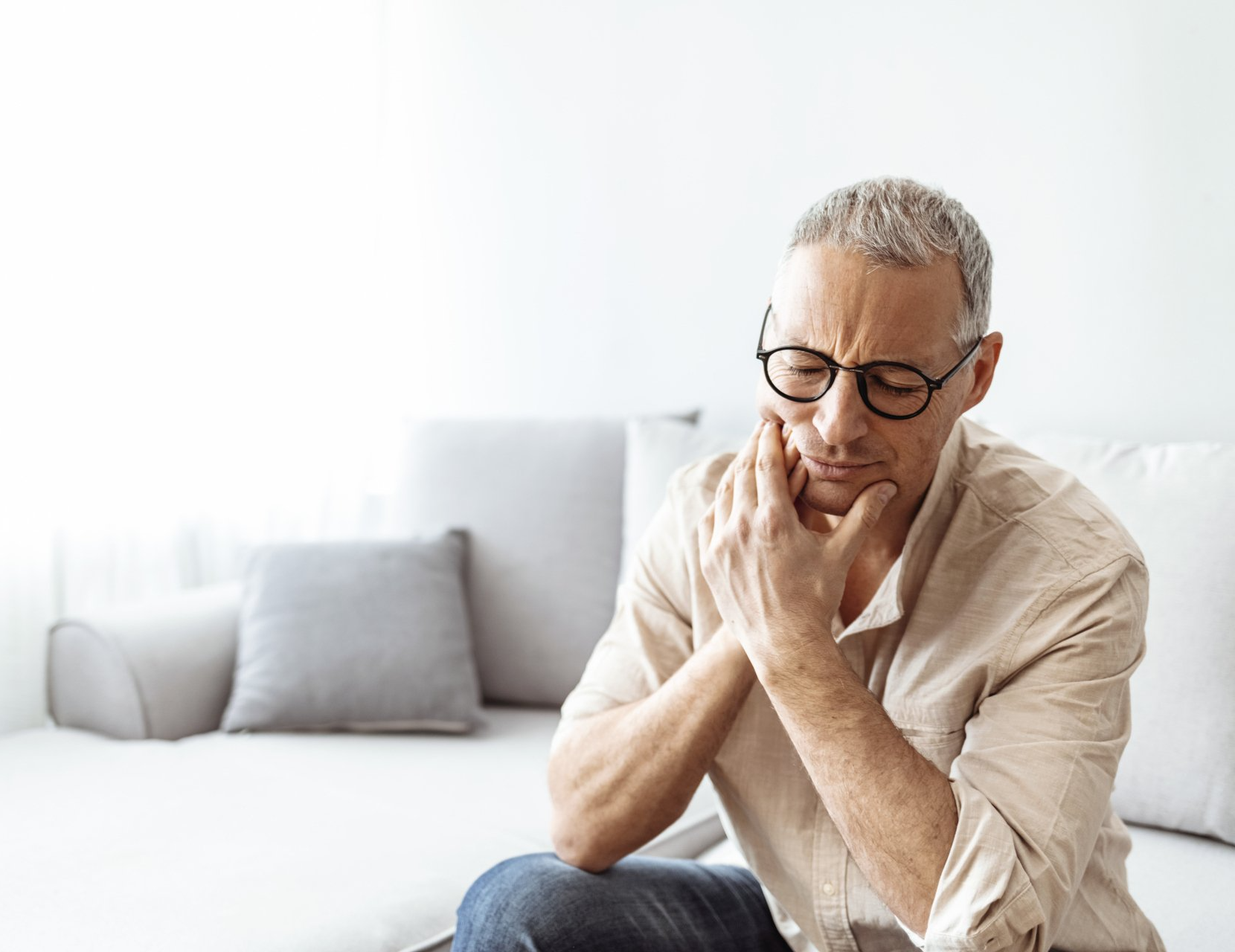 Man sitting on a couch, holding his face with a worried expression in a bright living room.