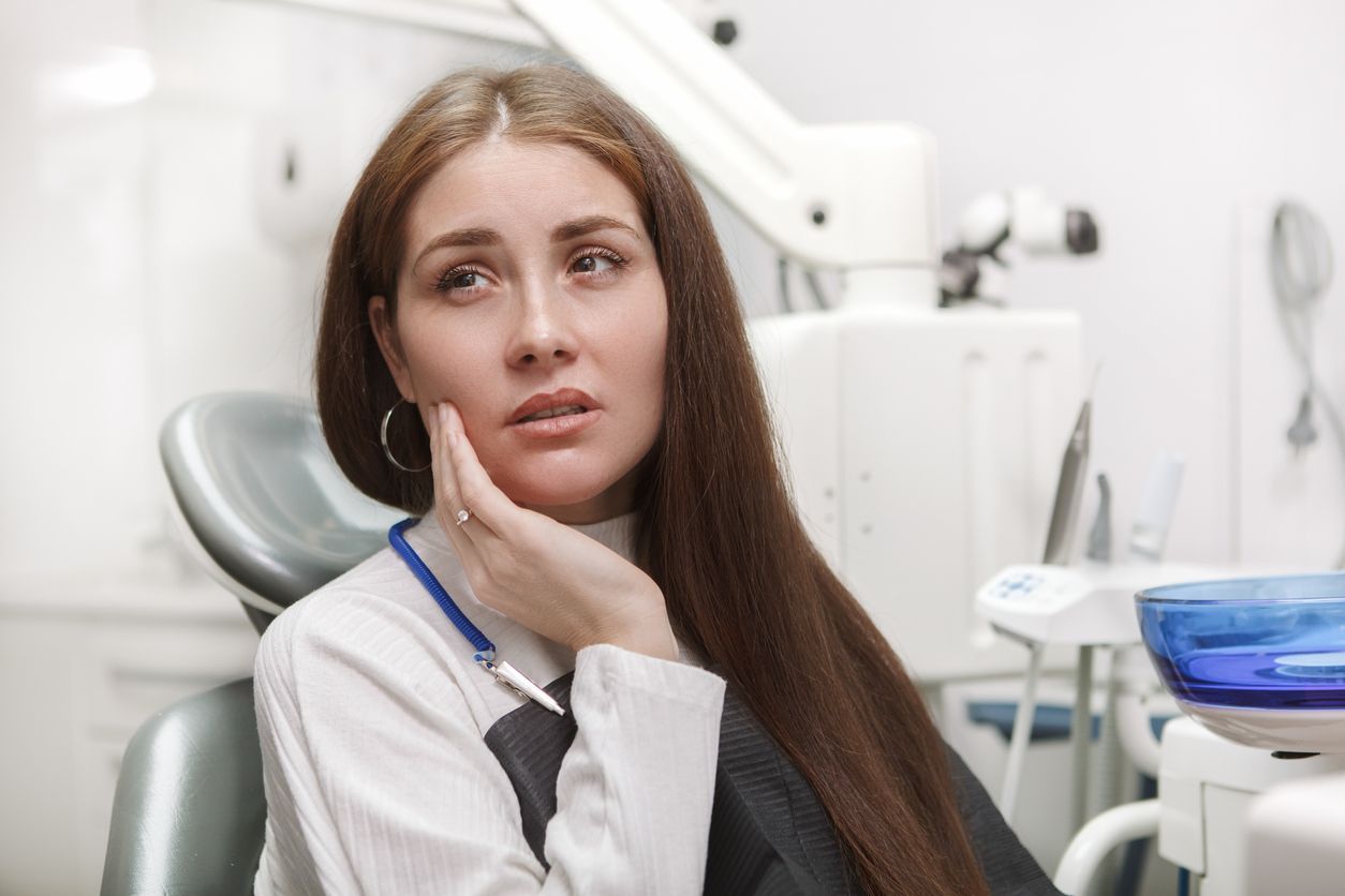 Woman in dental chair, holding jaw, appearing to be in pain, dental office setting.