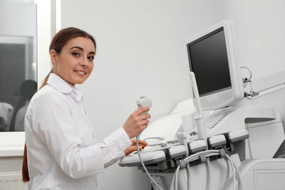 Woman holding ultrasound probe, standing near the machine in a clinic setting.