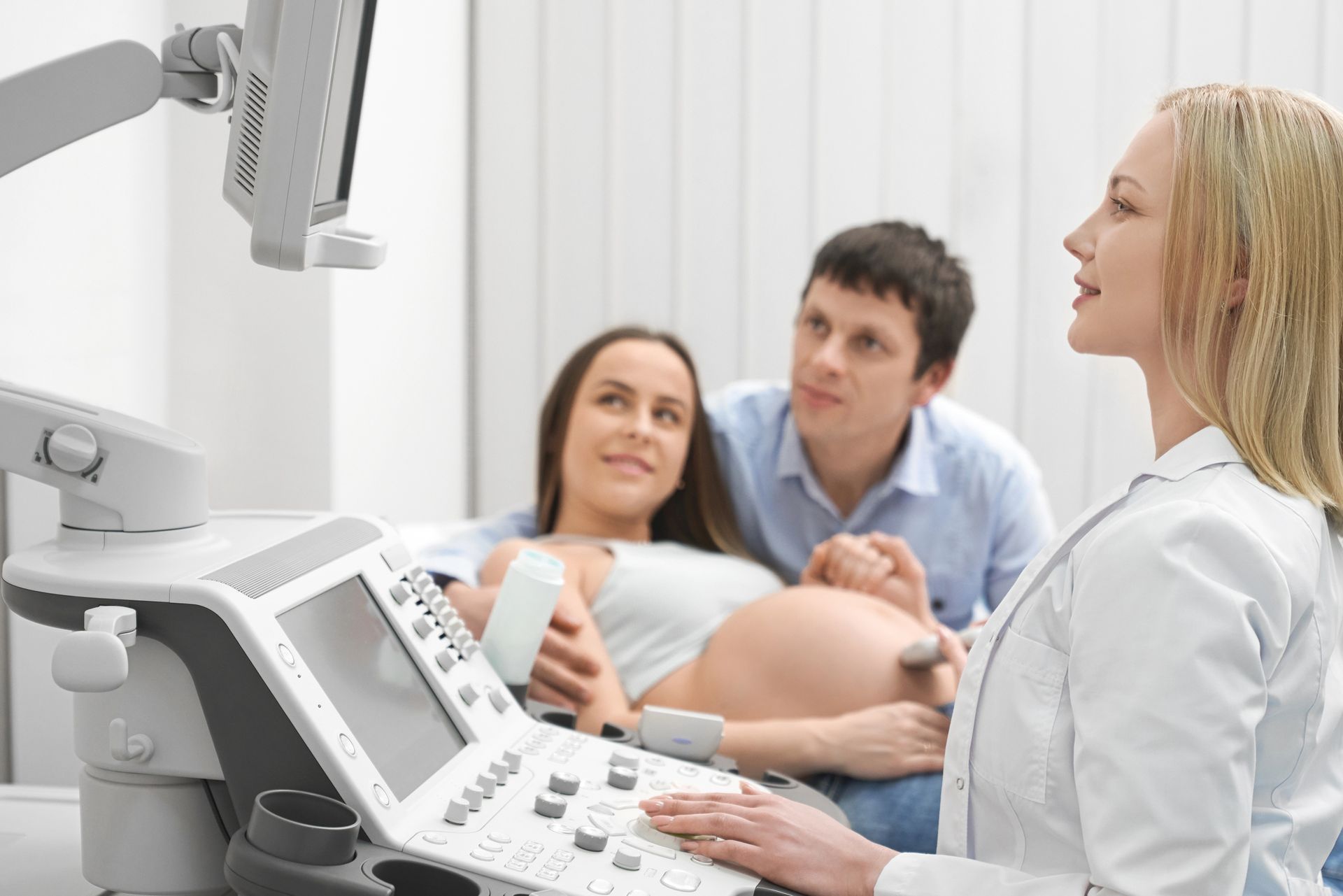 A medical professional performs an ultrasound on a pregnant patient while her partner watches from behind.