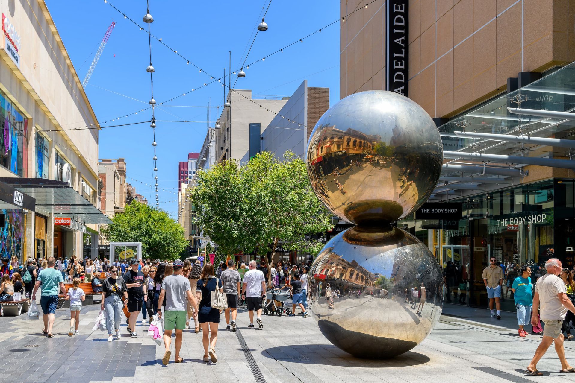 Pedestrian shopping street with crowds and a large mirrored silver sculpture under a blue sky