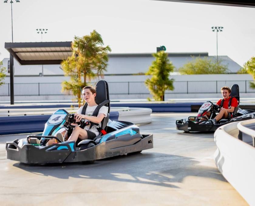 Two people driving blue-and-black go-karts on an outdoor track in bright sunlight