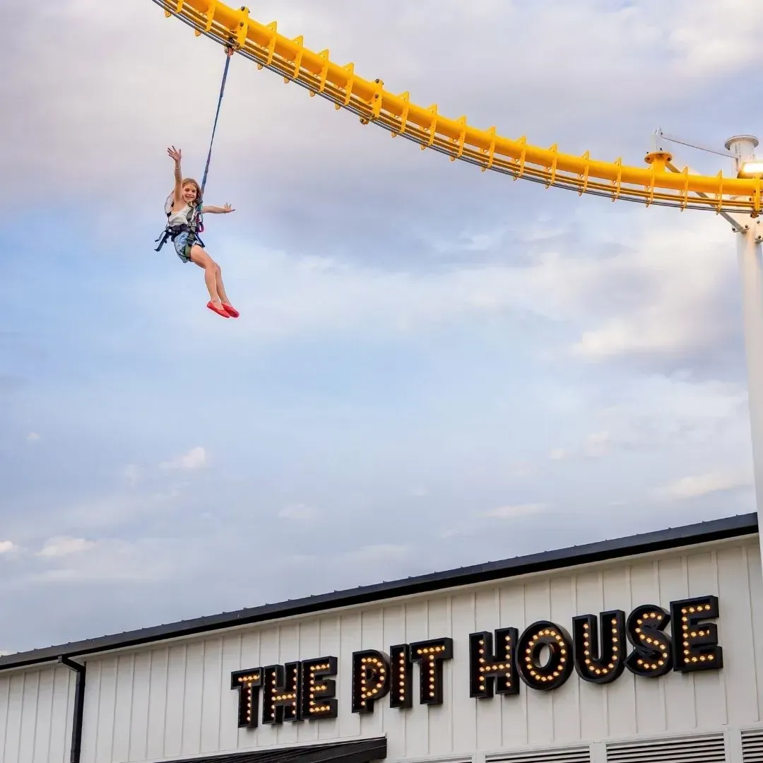 Person dangling from ropes course ride above a building labeled “THE PIT HOUSE”
