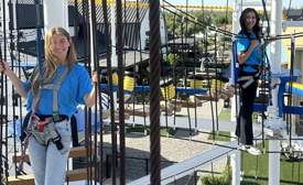 Two people in blue shirts use harnesses on a ropes course, standing on elevated platforms outdoors.