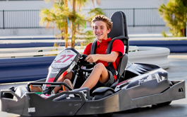 Person smiling in a black go-kart, driving outdoors on a sunny track.