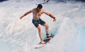 Surfer riding a wave in a blue surf pool, balancing on a board with arms outstretched.