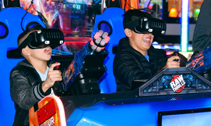 Two people wearing VR headsets play an arcade racing game in blue-lit cars.