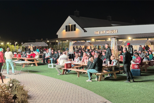 Crowded outdoor evening gathering at a lit house with people seated at picnic tables on a patio and lawn