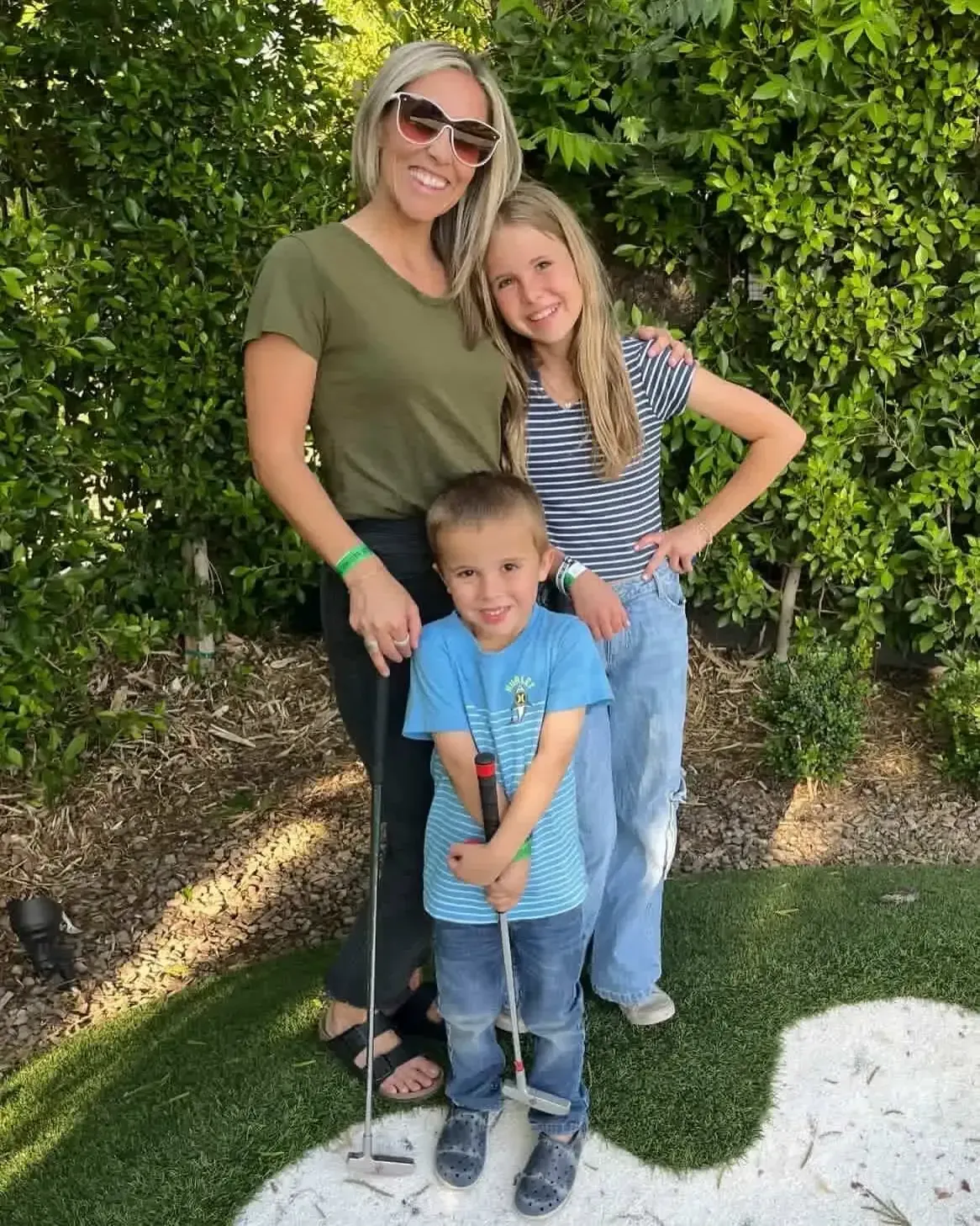 Three people posing outdoors by a green hedge, smiling, with a child holding a golf club.