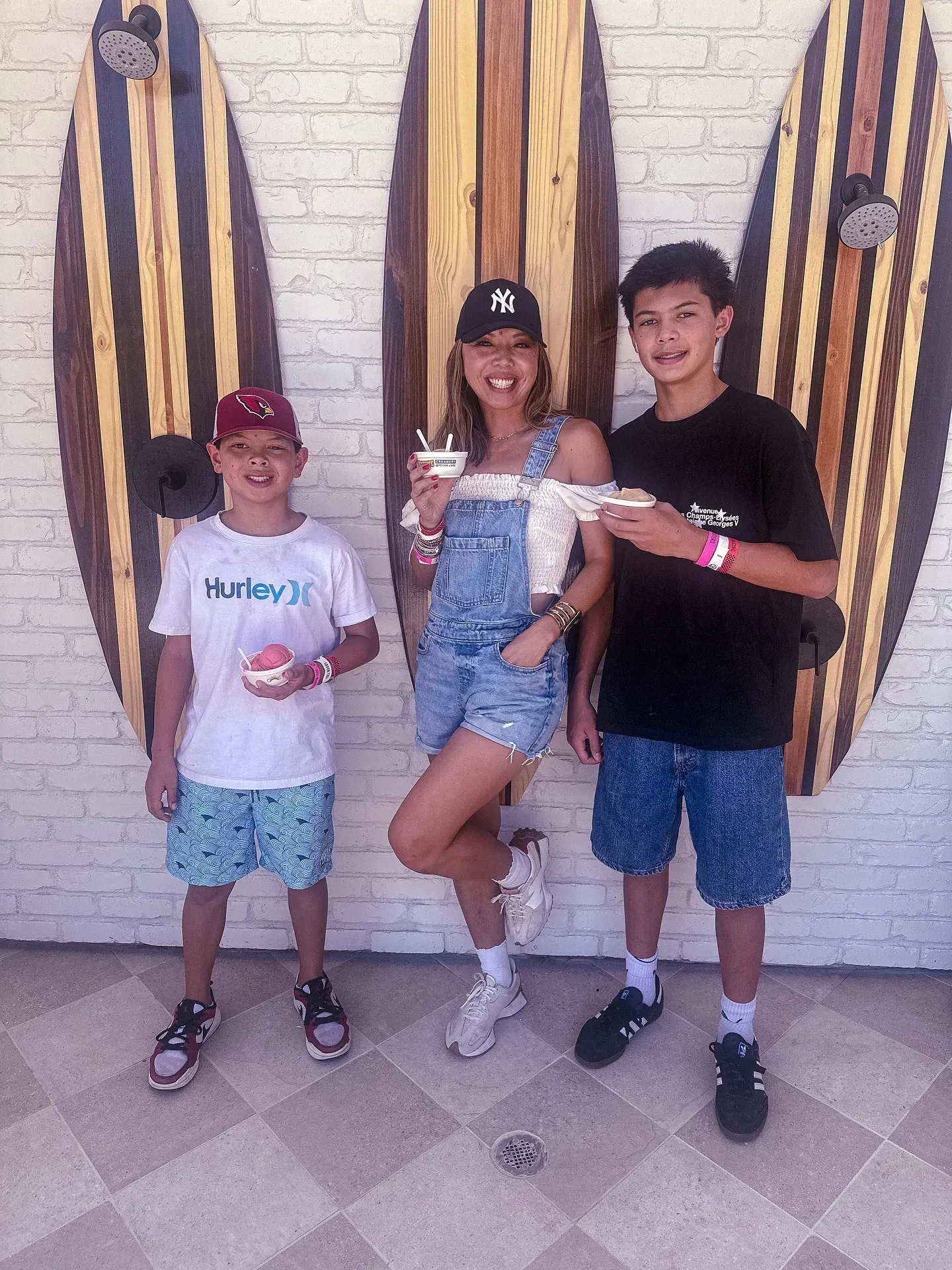 Three people posing outdoors in front of surfboards and a brick wall, smiling and holding ice cream.