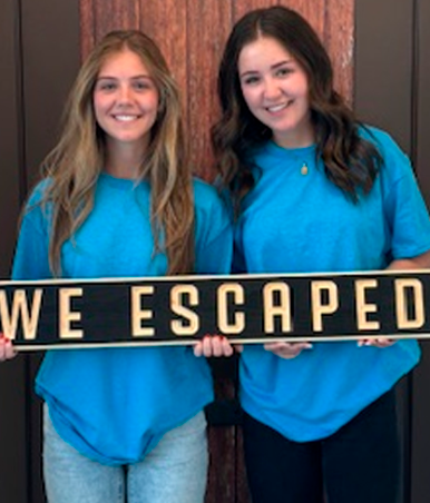 Two smiling women in blue shirts holding a sign that says “WE ESCAPED”