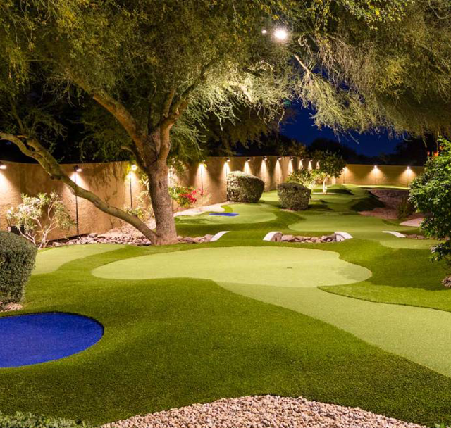 Nighttime mini golf course with bright green putting greens, blue sand traps, and lit trees