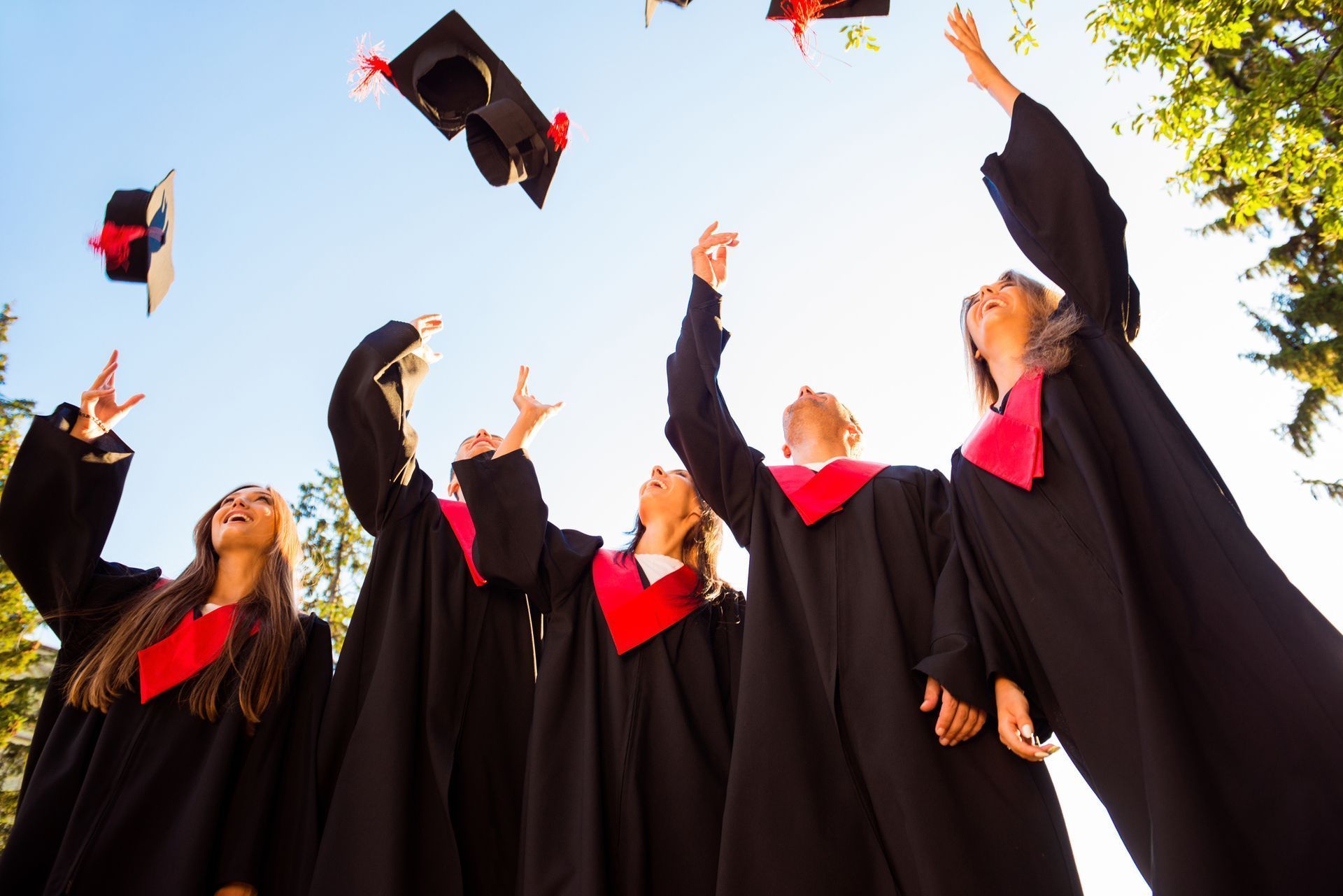 Graduates in black gowns and red stoles toss caps into the air against a bright sky