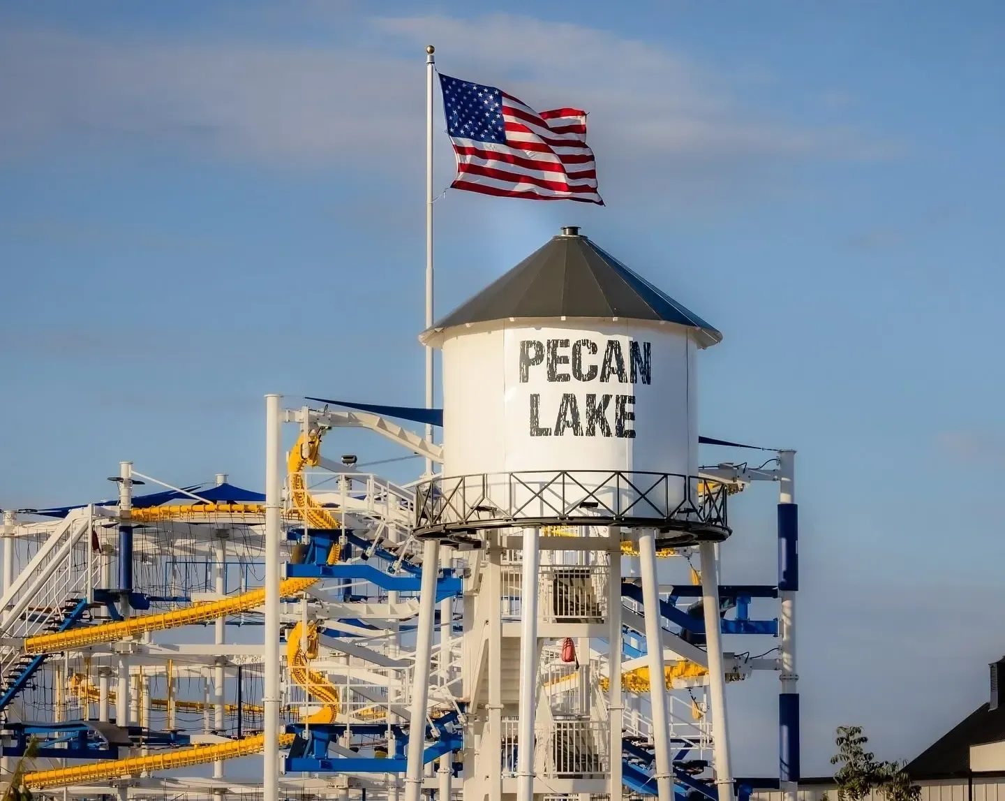 Pecan Lake water tower with American flag and amusement park rides under a blue sky