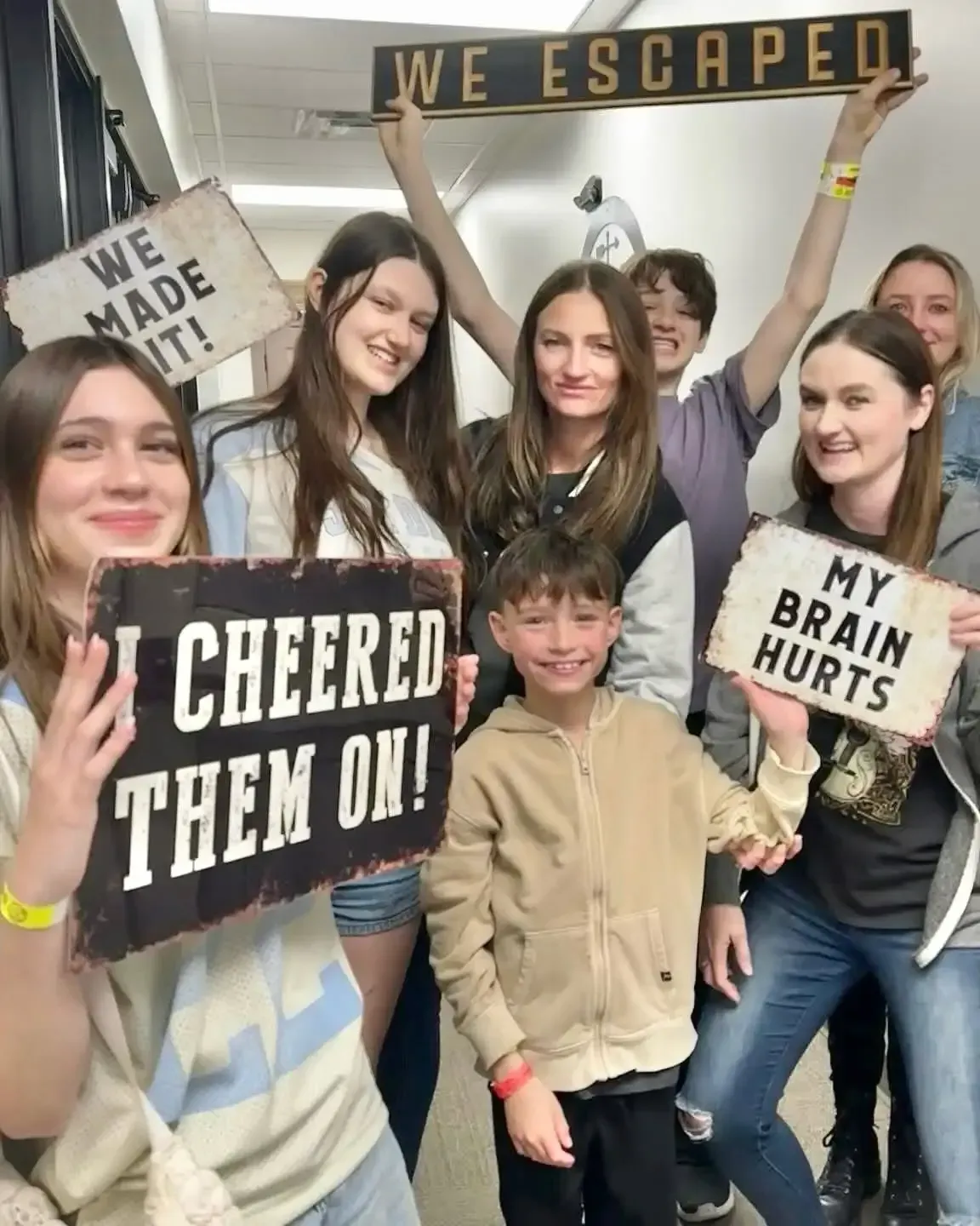 Group of smiling people holding escape room signs: “WE ESCAPED,” “WE MADE IT,” “I CHEERED THEM ON!,” and “MY BRAIN HURTS”