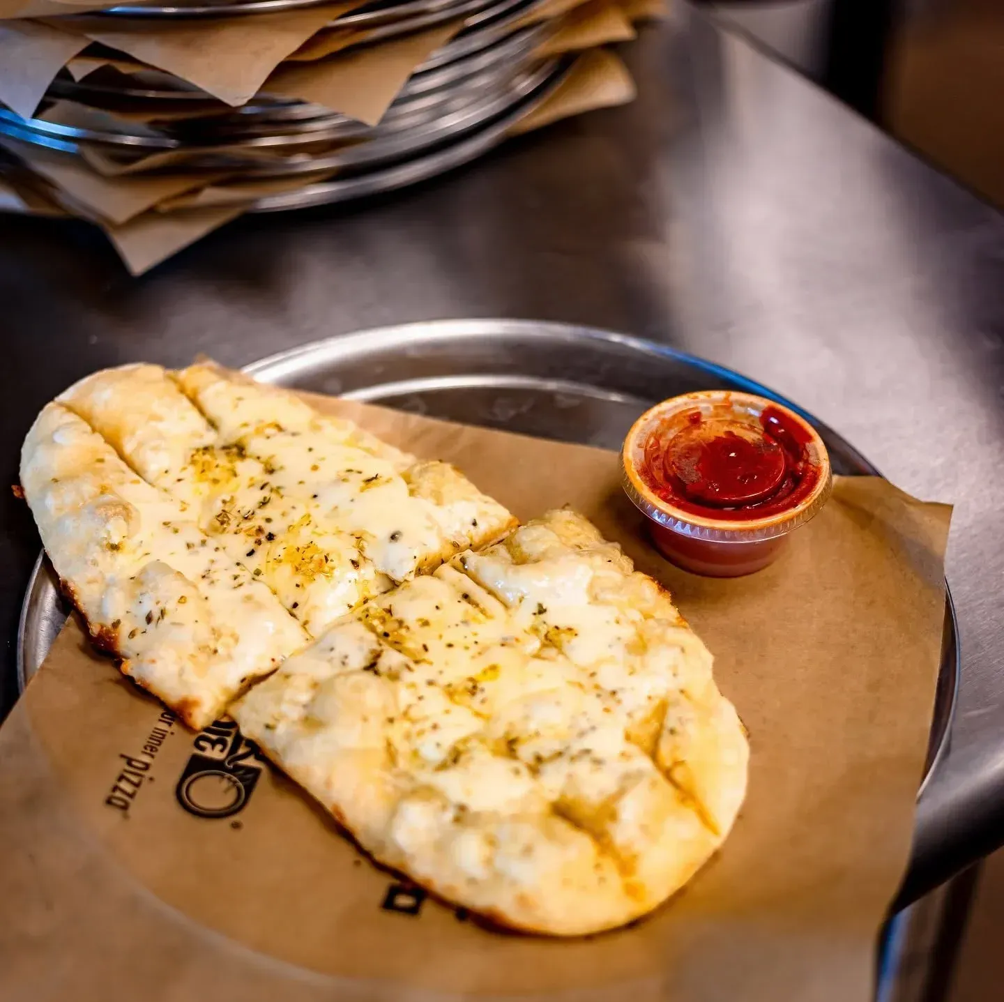 Cheesy flatbread on brown paper with a small cup of red dipping sauce on a metal table