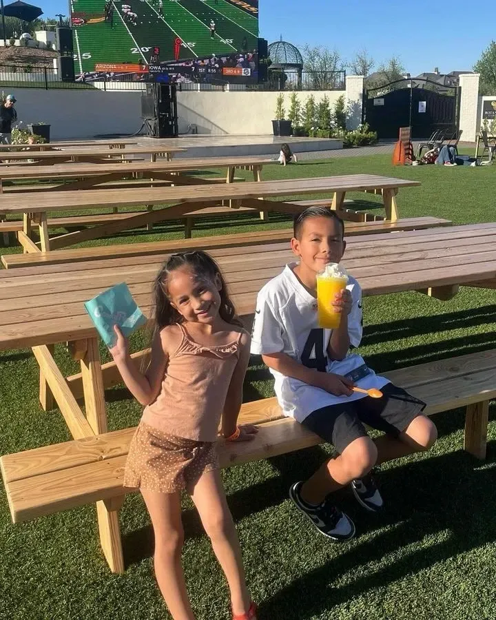 Two children sit on a picnic table bench outdoors, smiling and holding colorful drinks at a stadium.