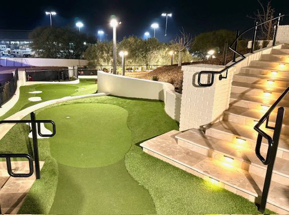 Nighttime outdoor mini golf course with green turf, lit stairs, and black handrails