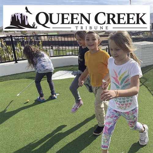 Children playing on green turf in front of a Queen Creek Tribune sign.