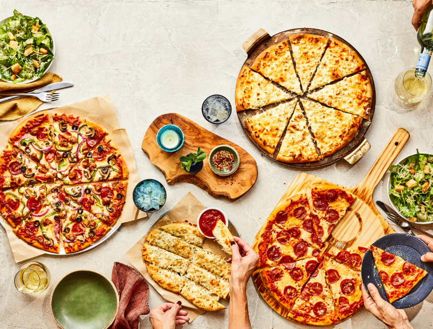 Overhead spread of assorted pizzas on a table with plates, drinks, and serving utensils