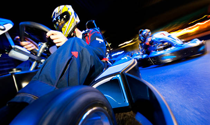Go-kart racers speeding through a blue-lit track, one driver close to the camera in a yellow helmet