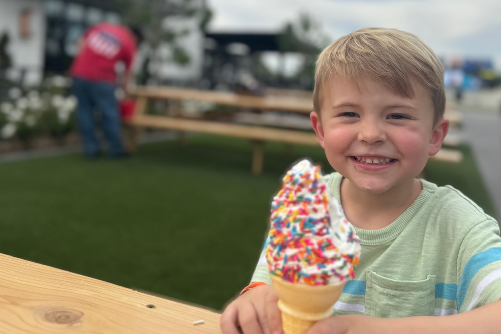 Smiling child holding a rainbow-sprinkled ice cream cone at an outdoor table