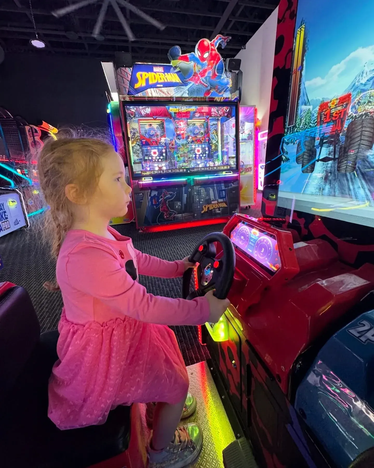 Child in a pink dress playing a red arcade driving game in a brightly lit arcade