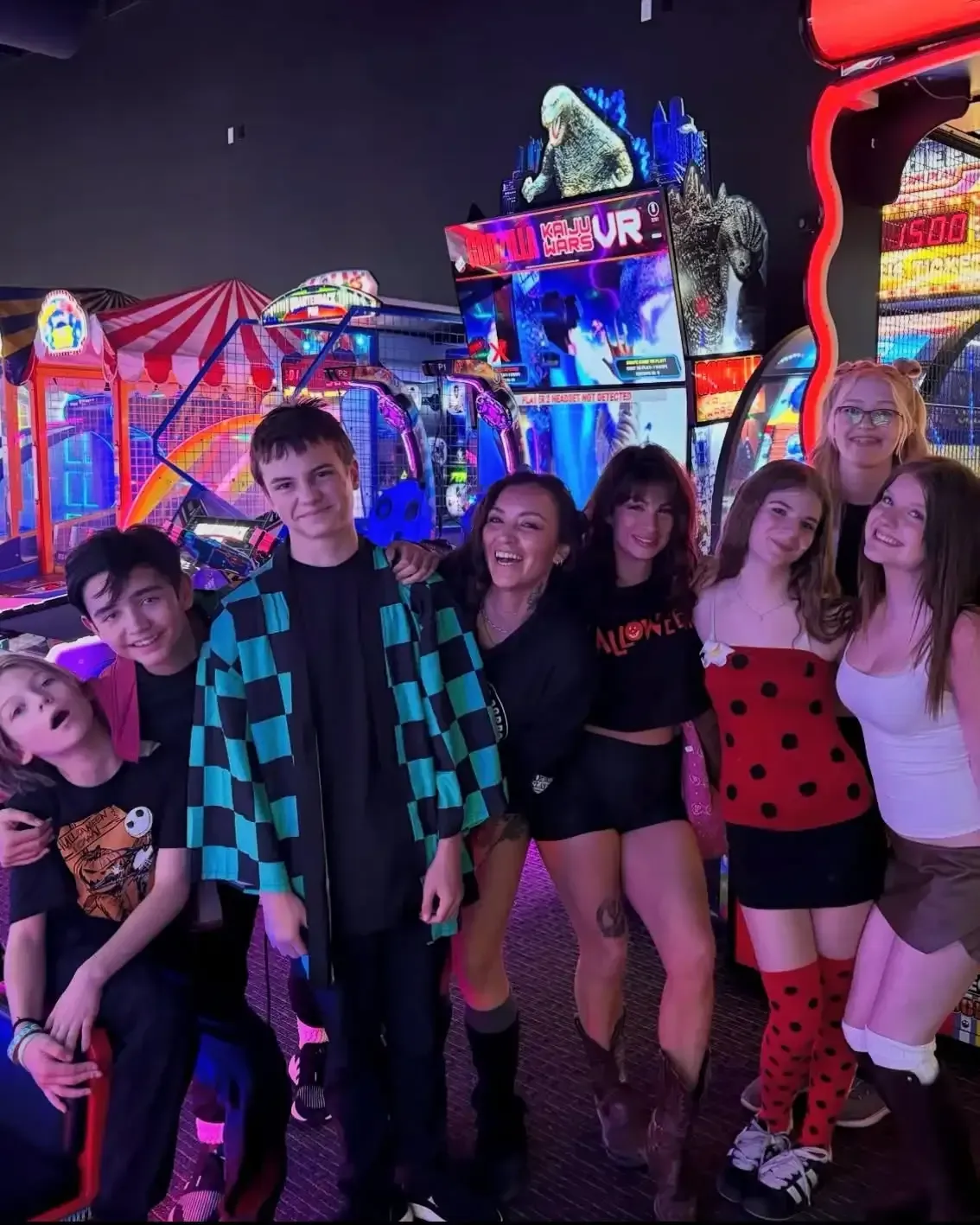 Group of people posing in front of glowing arcade machines, wearing colorful outfits and smiling.