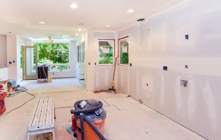 Renovated empty room with white walls, tools on the floor, and large windows opening to greenery