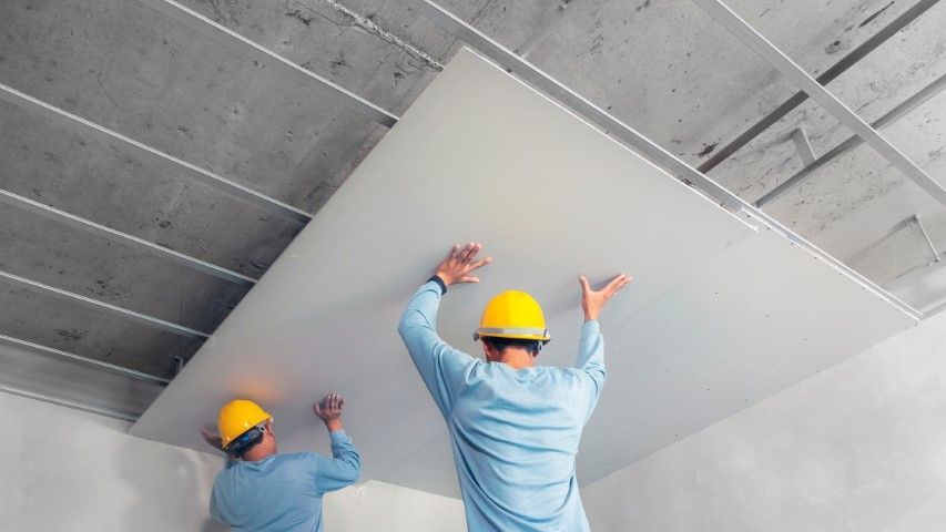 Two workers in yellow hard hats install a large white ceiling panel indoors.