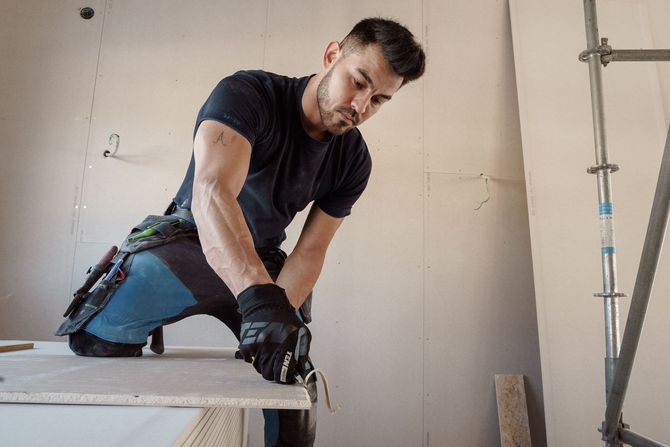 Worker kneeling on a countertop, using a tool during construction in an unfinished room.