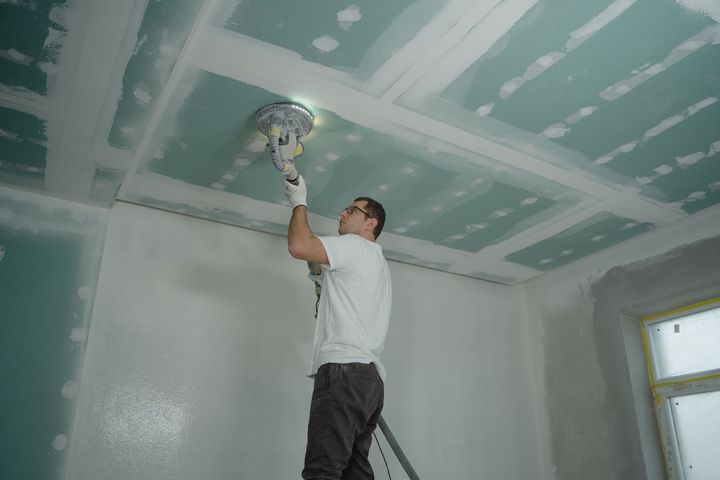 Person sanding a green drywall ceiling with a power sander in a room under renovation