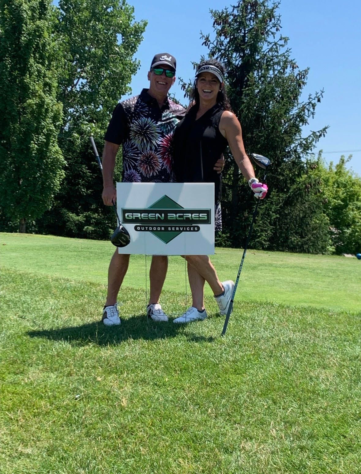 Man and woman smiling, posing with golf clubs and a sign that says
