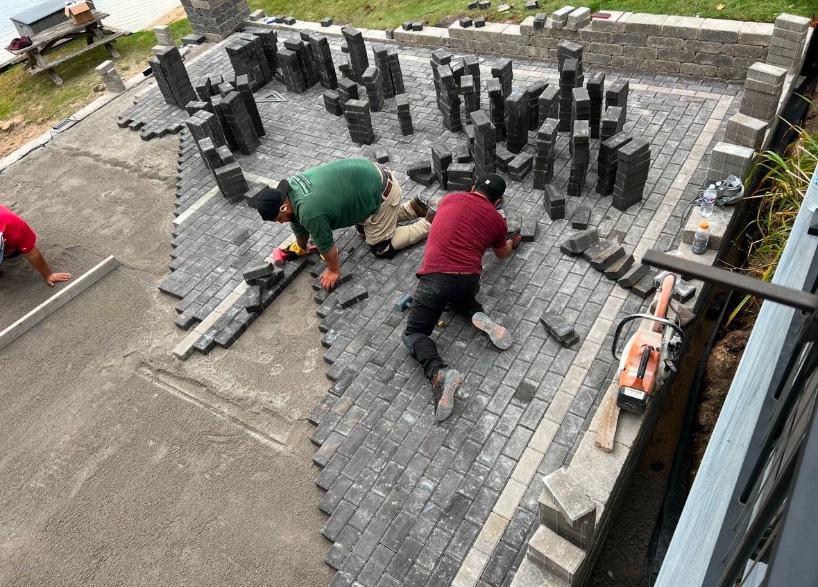 Workers laying dark gray cobblestones on a patio near a wall and a lake.