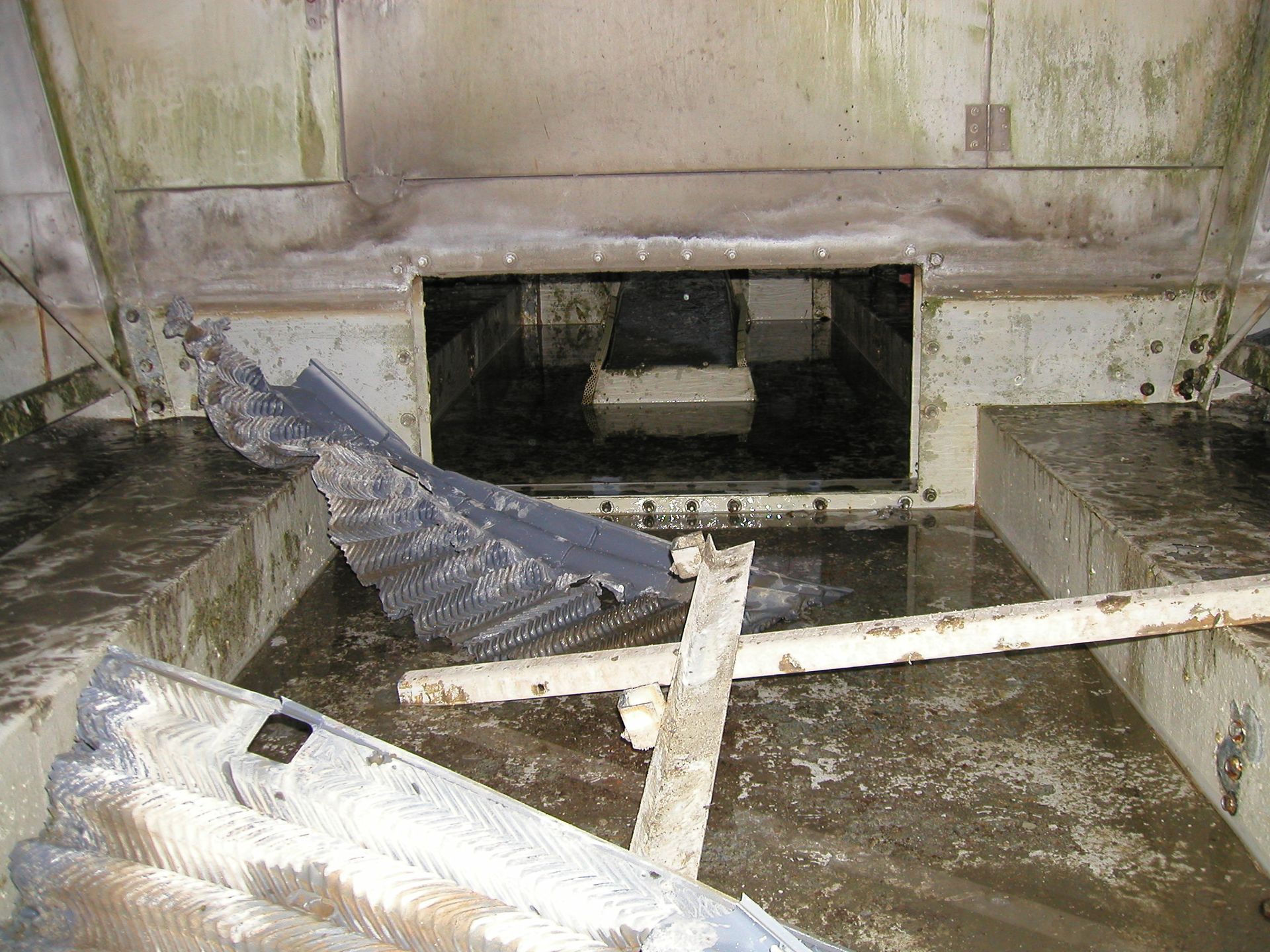 Damaged flooded industrial room with debris, broken equipment, and a large open tank opening