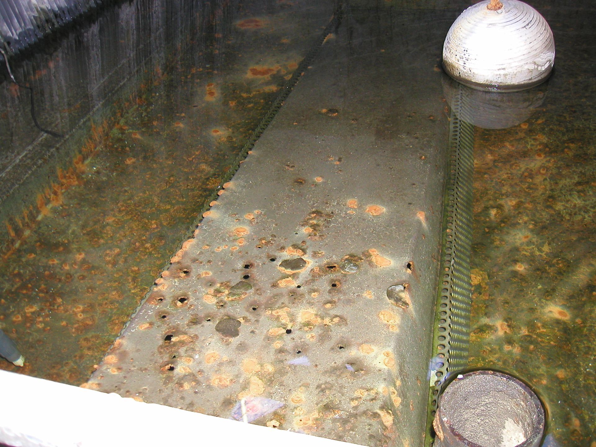 Dirty flooded concrete trench with debris, a white buoy-like float, and a metal grate at the end