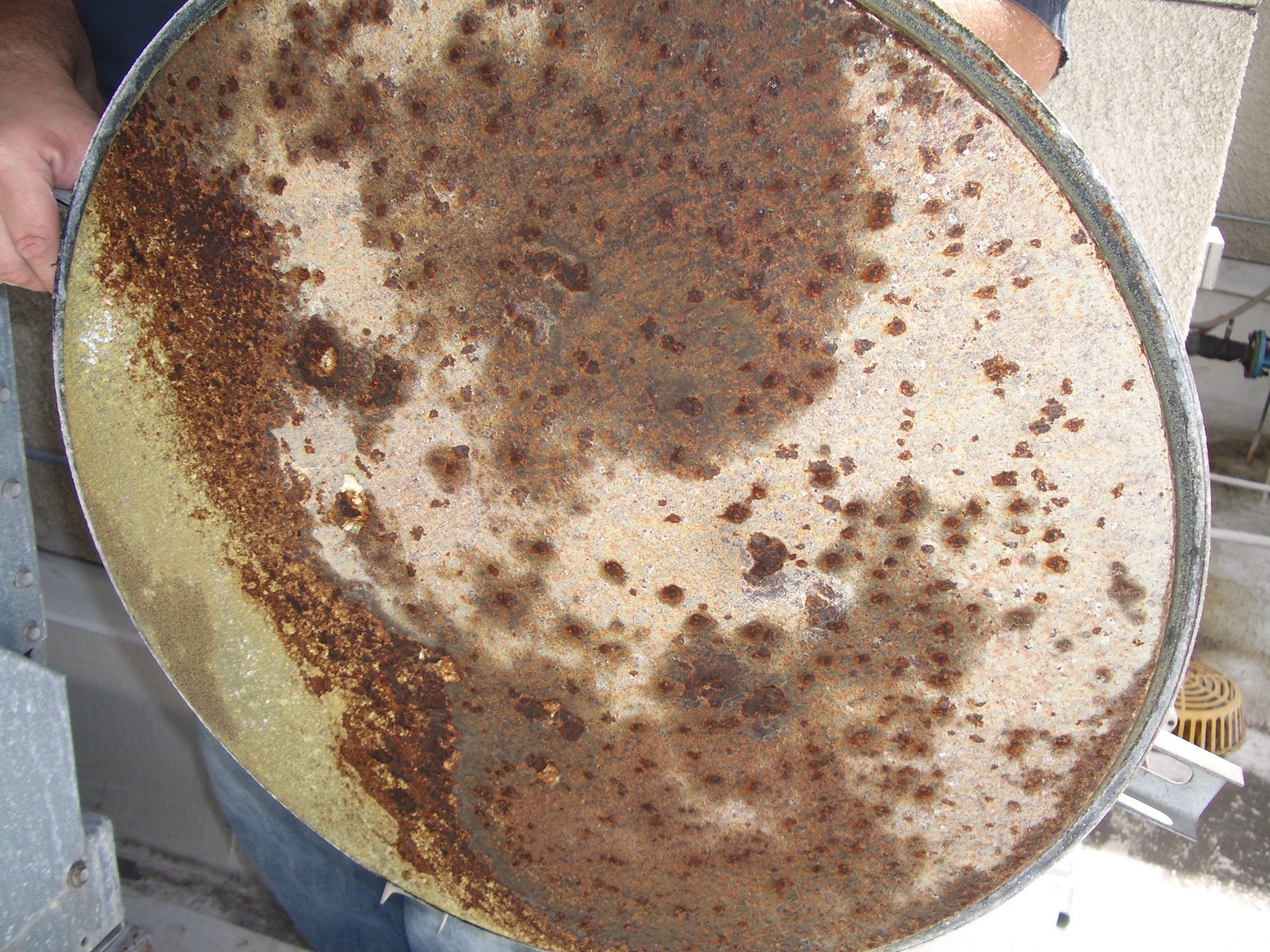 Rusty, weathered round metal dish with heavy brown corrosion and stains, held outdoors by a person’s hand