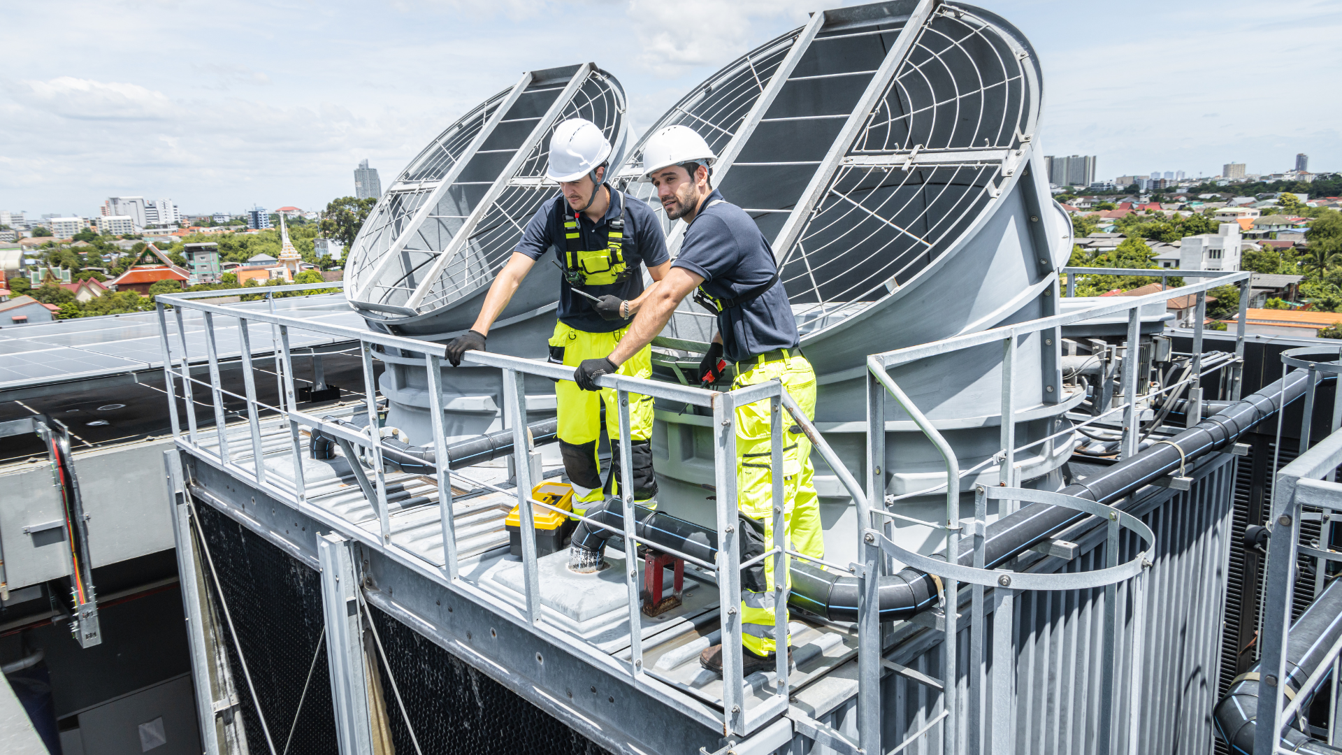 Two workers in helmets on a rooftop beside large industrial fans and metal railings
