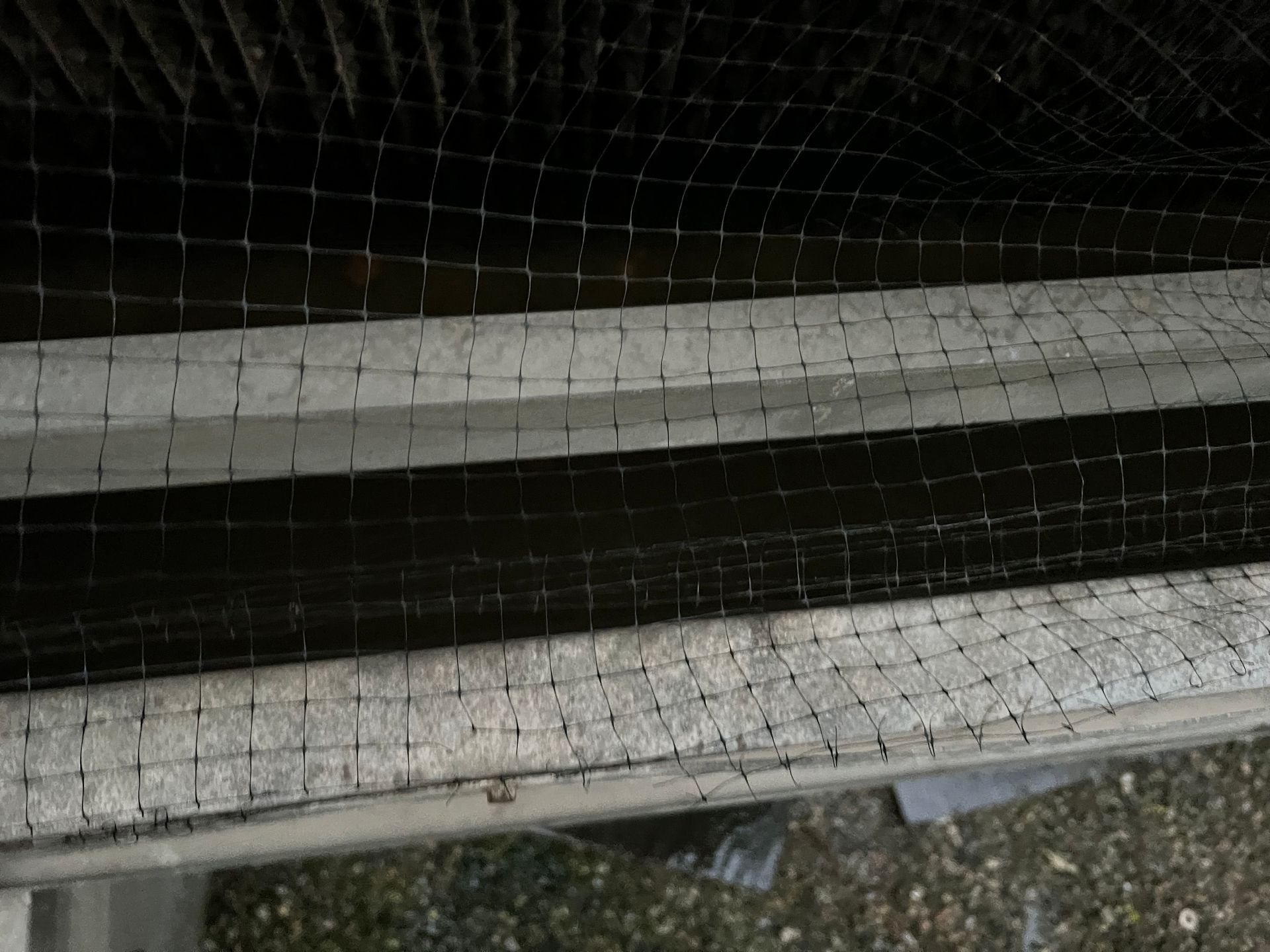 Close-up of a dirty metal railing with fine netting in front of dark background and gravel below